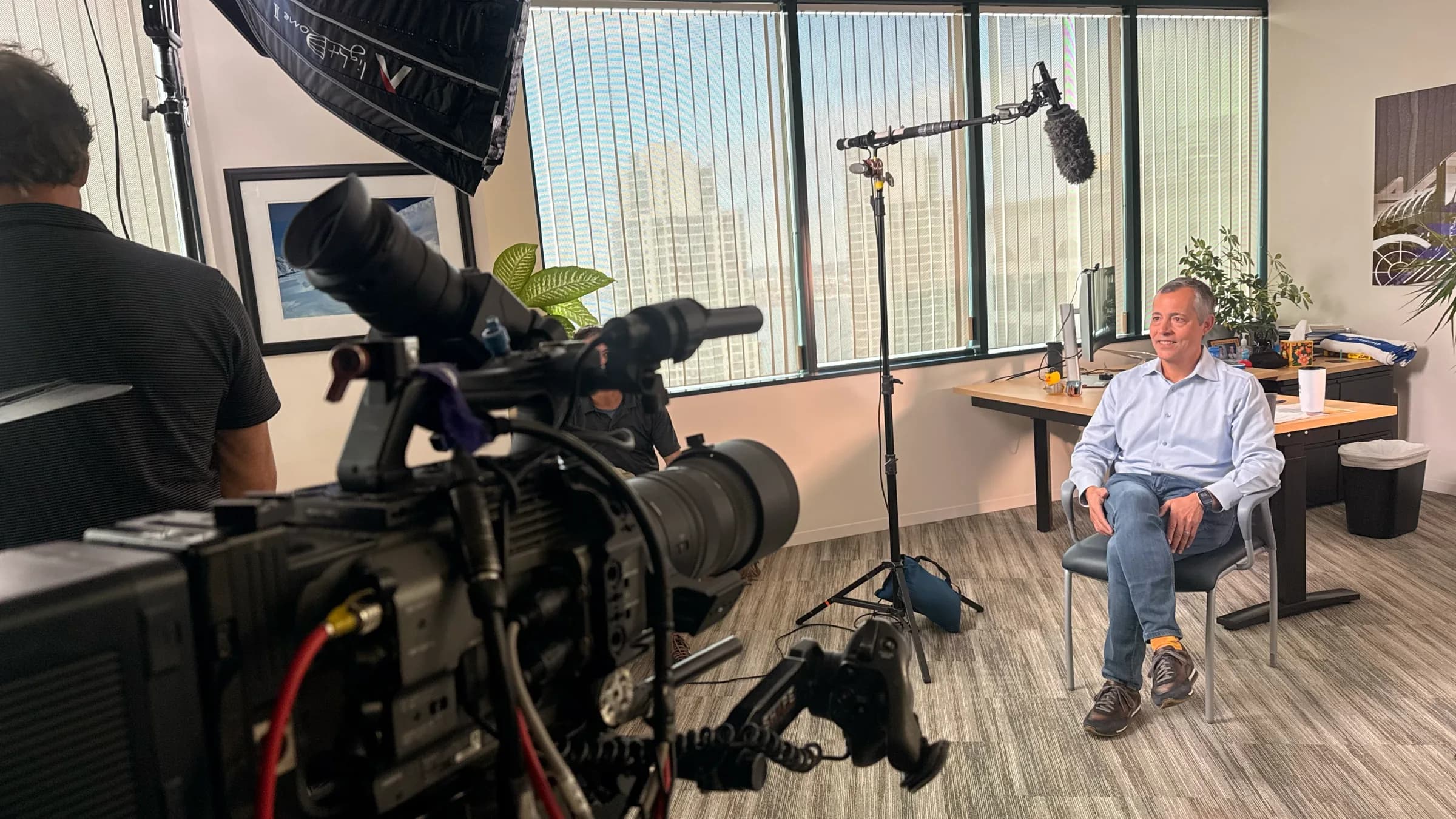 Interview setup in an office with a cinema camera close in the foreground, a boom mic, and a seated subject by city windows