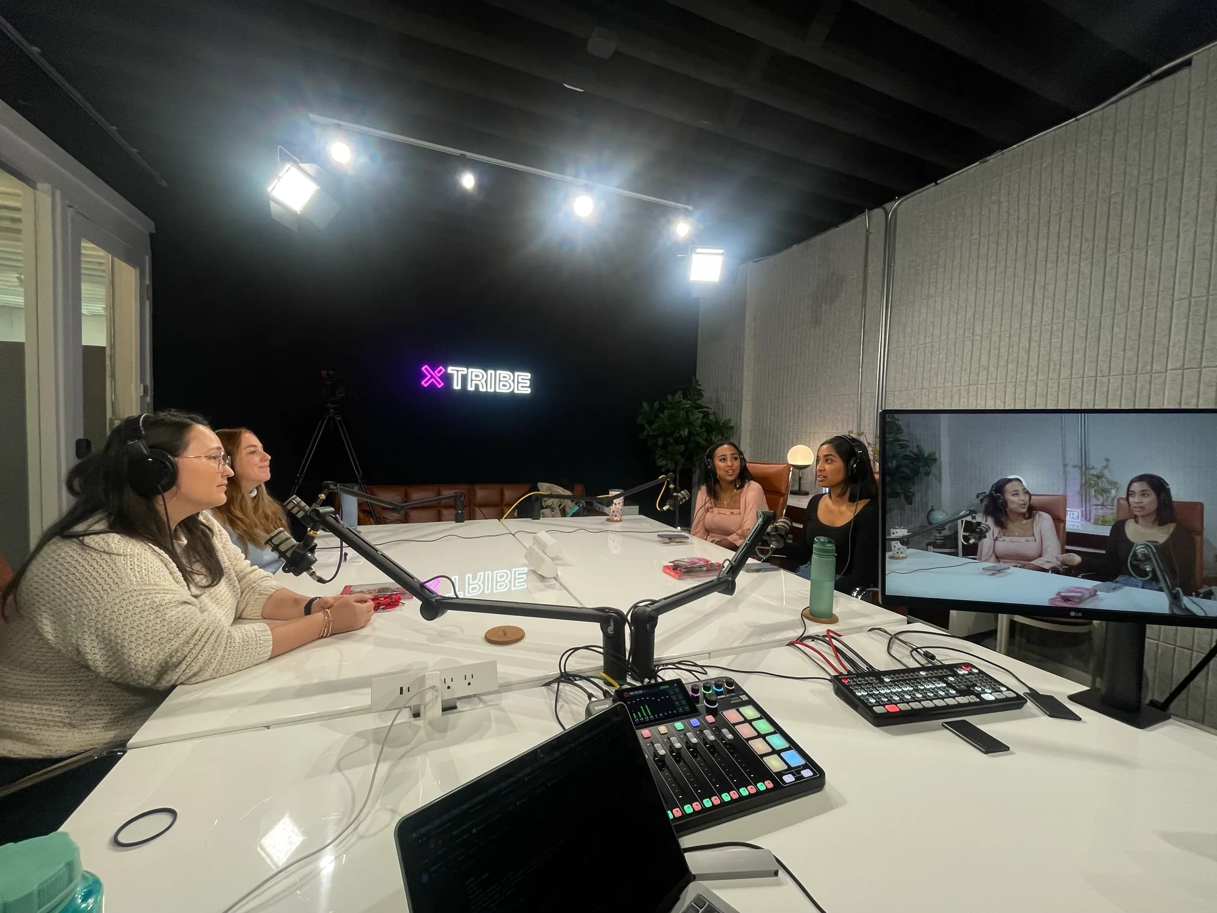 Four podcast hosts in headphones at a white table with boom mics, a mixer, and a monitor showing the feed